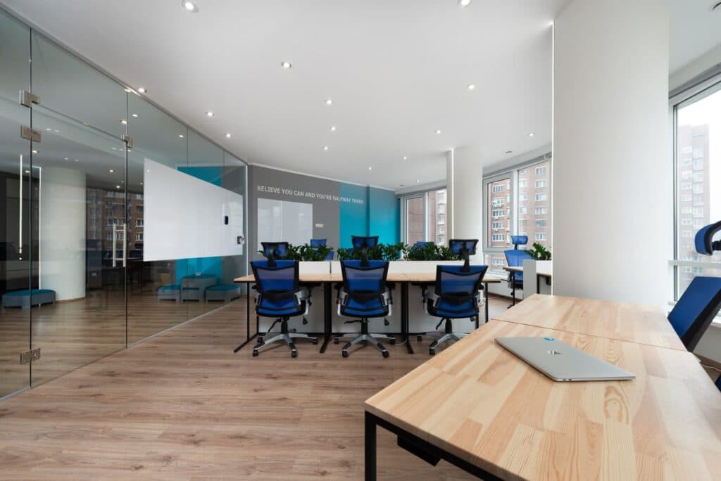 Office desks and blue chairs beside a glass partition with a hinged door and a broad white privacy band.