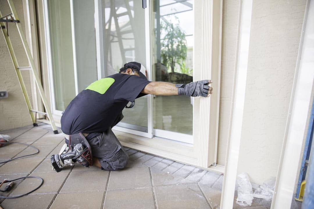 A glazier kneels on a patio and fits the frame of a sliding glass door.