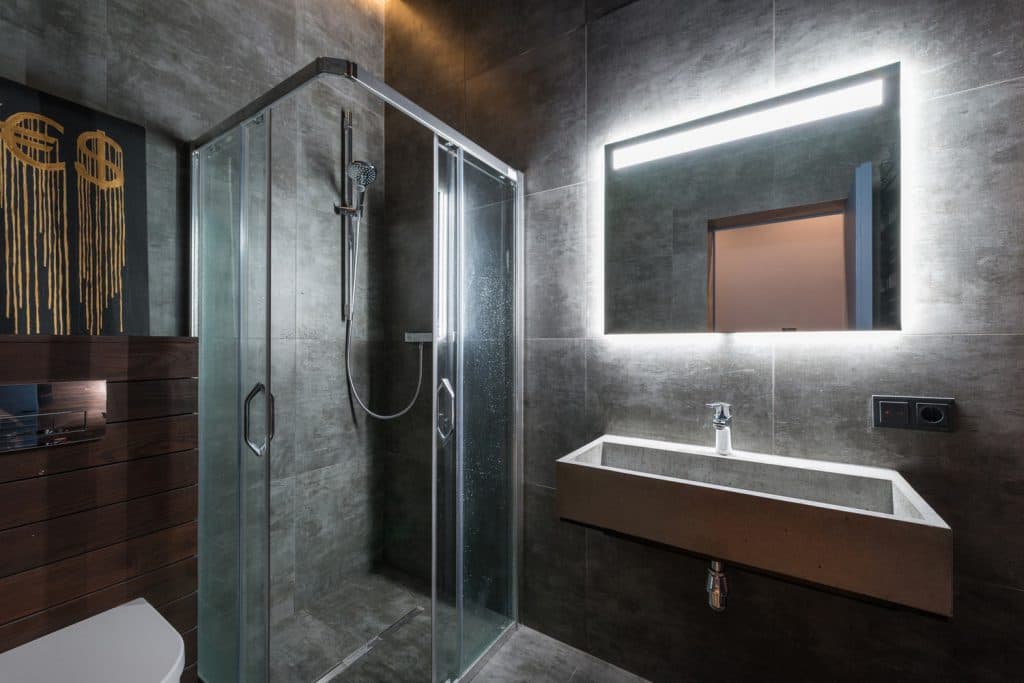 Glass shower enclosure with metal framing beside a wall-mounted basin in a grey tiled bathroom.