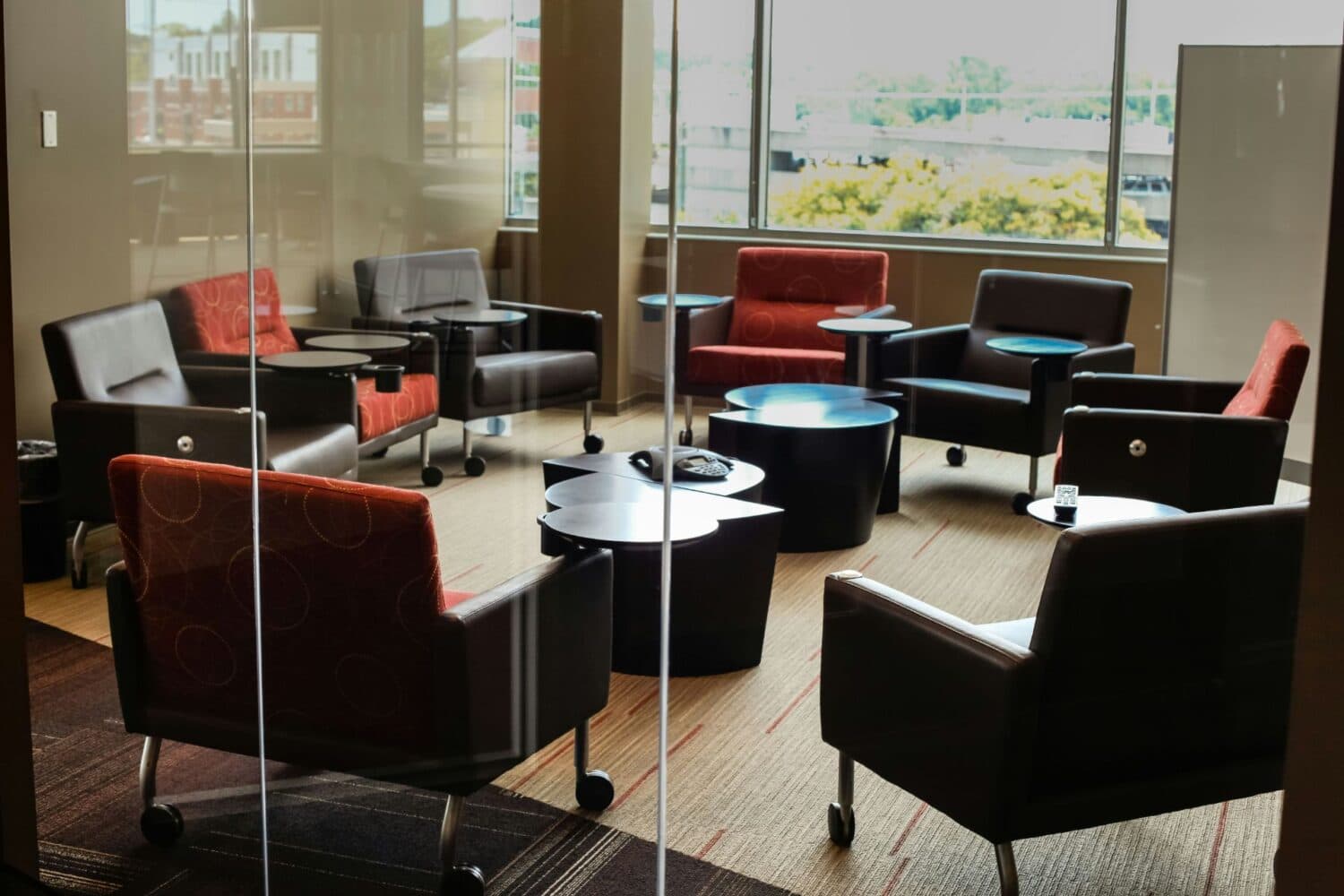 Red and dark lounge chairs and small tables seen through an office glass partition, with windows beyond.